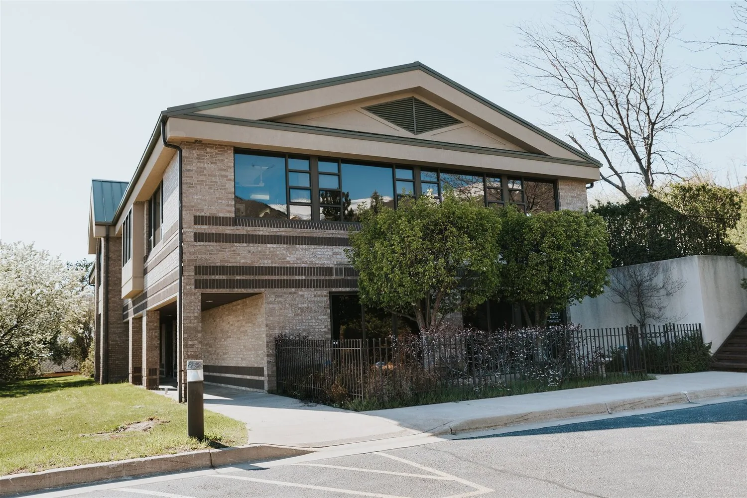 Exterior of a two-story brick office building with large front windows, landscaping trees, and an adjacent sidewalk.