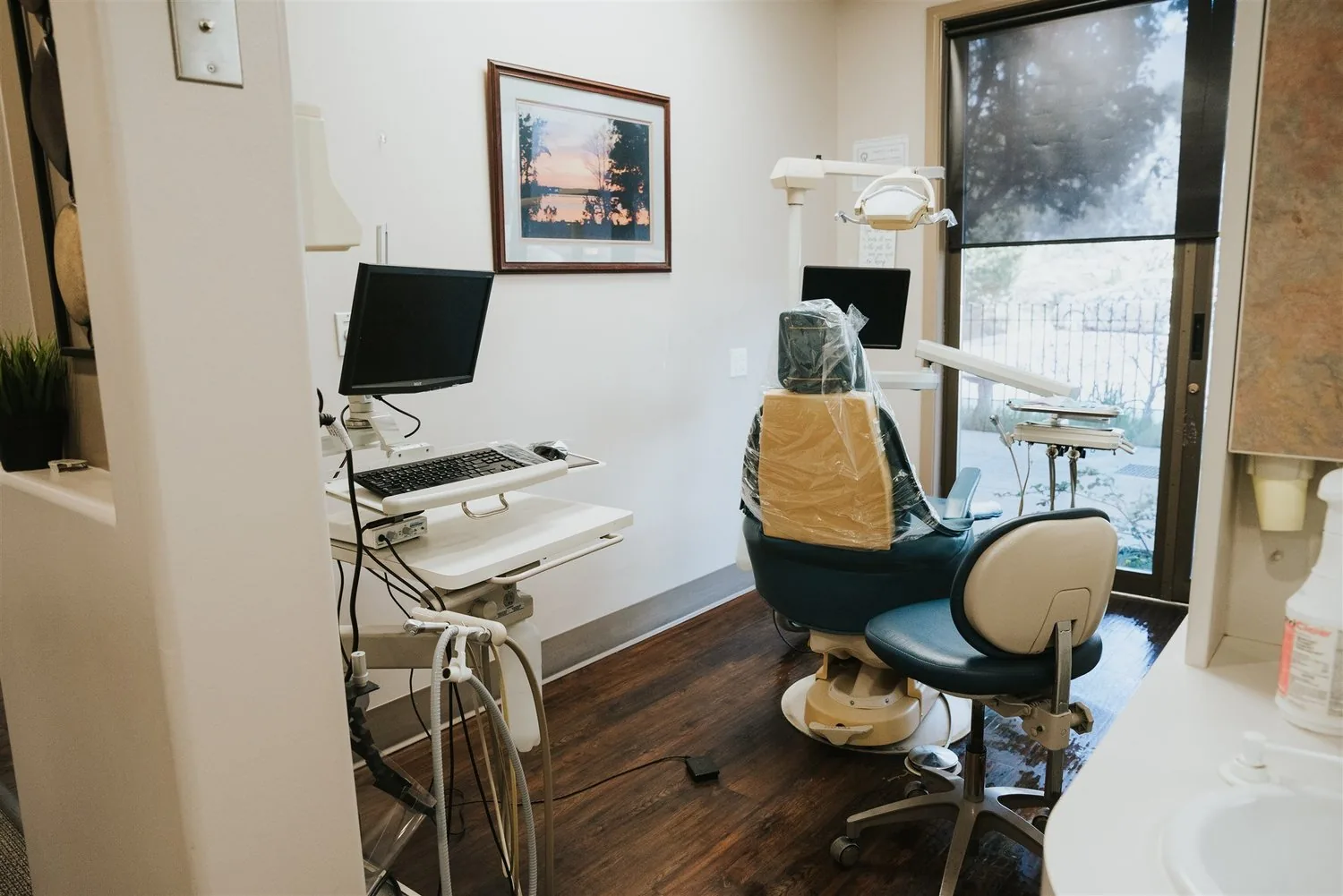 Dental treatment room with an exam chair, computer station, and dental equipment by a large window.
